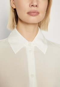 Camicia bianca con colletto a punta, caratterizzata da una superficie liscia e bottoni trasparenti. Primo piano del colletto e della parte superiore del petto.