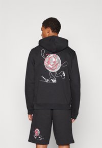 Svart hoodie i sweatshirtmaterial med en grafisk design av en leende tecknad karaktär i rött och vitt på ryggen.
