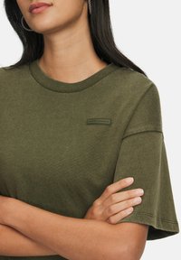 Nainen, jolla on pitkät tummat hiukset, pukeutuneena oliivinvihreään Under Armour -teeshirtiin, kädet ristissä, renkaita korvakoruina, valkoista taustaa vasten.