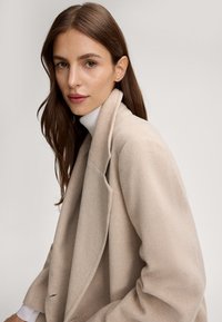 Beige wollen jas met een geknipt revers, losse pasvorm en dubbelzijdige sluiting. Hoog hals wit topje zichtbaar daaronder.