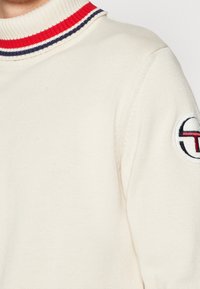 Cremefarbener Pullover mit kragen aus rot-blau gestreiftem Rippstrick, versehen mit einem runden Logo-Patch am linken Ärmel. Gestrickte Textur.