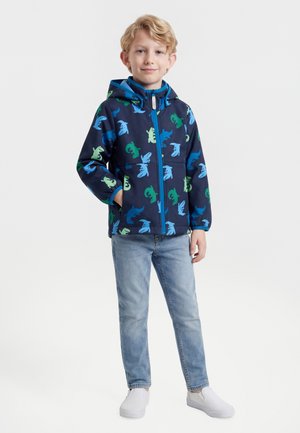 Un garçon blond se tient debout en souriant, portant une veste à capuche bleu marine avec un imprimé dinosaure, un jean bleu clair et des chaussures blanches sans lacets, sur un fond uni.