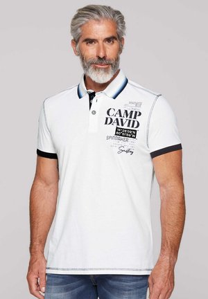 Camisa polo blanca con cuello azul y negro, que presenta un gráfico de "CAMP DAVID" y coordenadas náuticas. Material de algodón, mangas cortas.