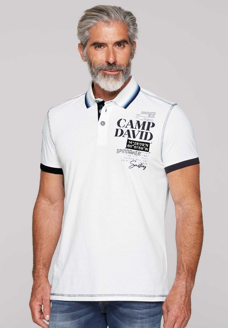 Polo bianco con colletto blu e nero, con grafica "CAMP DAVID" e coordinate nautiche. Materiale in cotone, maniche corte.