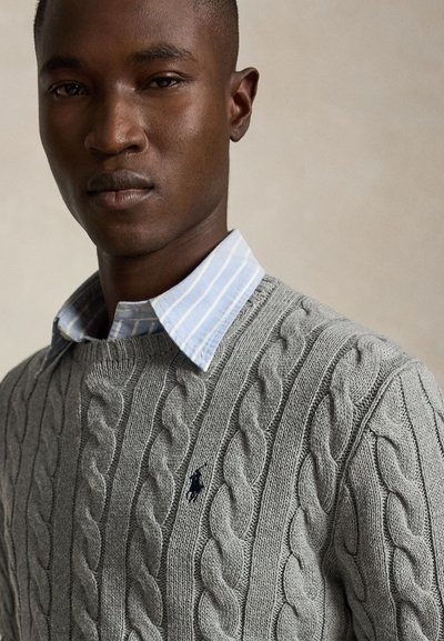 Uomo che indossa un maglione grigio a trecce sopra una camicia a righe blu e bianca con colletto, su sfondo neutro.