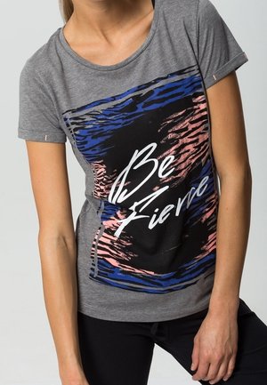 Kobieta nosząca szarą koszulkę z niebieskim, czarnym i różowym abstrakcyjnym wzorem oraz białym napisem "Bądź zawzięta."