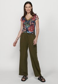 Camisa de cuello en V con estampado floral en azul, rojo y dorado, combinada con pantalones sueltos en verde oliva que cuentan con bolsillos laterales y una cinturilla elástica.