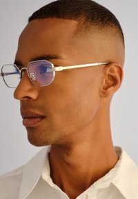 Νεαρός άνδρας με κοντά μαλλιά που φοράει ασημί γυαλιά με εξαγωνικό σκελετό και λευκό πουκάμισο με γιακά, κοιτάζει προς τα αριστερά σε γκριζό φόντο.