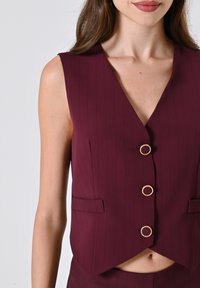 Gilet bordeaux senza maniche con scollatura a V, motivo a righe, due tasche frontali e tre bottoni circolari dorati. Tessuto testurizzato.