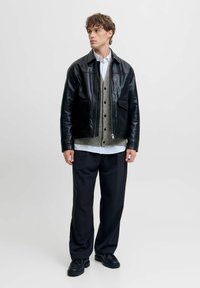 Schwarze Lederbomberjacke über einem grauen Cardigan mit Knöpfen, darunter ein weißes Hemd und dunkle Hosen, kombiniert mit schwarzen Schnürschuhen.