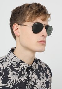 Ochelari de soare Ray-Ban aviator cu lentile întunecate, cadru subțire din metal negru, purtați de un model îmbrăcat într-o cămașă cu model de frunze în culori negre și crem.