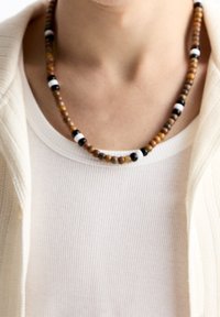 Osoba nosící náhrdelník z hnědých, černých a bílých korálků přes bílé žebrované tričko a krémový kardigan.