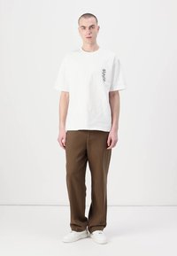 White cotton t-shirt with a black floral design on the left chest, paired with brown trousers and white sneakers. Simple, relaxed fit.