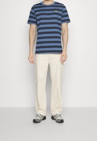 Man som bär en marinblå och blårandig t-shirt, off-white byxor och gråsvarta sneakers som står mot en vit bakgrund.