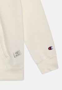 Hoodie di cotone crema con tasca laterale, polsini a costine e logo ricamato sulla manica. Presenta un'etichetta con la scritta "Sii il tuo campione".