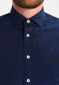 Hombre con barba que lleva una camisa de cuadros azul oscuro abotonada, con botones blancos, el cuello abrochado y un logotipo bordado en el pecho.