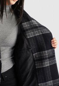 Femme portant un haut gris côtelé et un pantalon noir, ouvrant un manteau à carreaux noir et blanc pour montrer la doublure intérieure noire lisse avec poche.