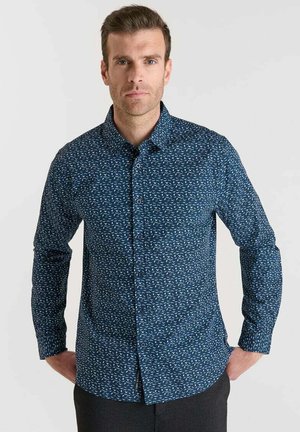 Man met kort bruin haar in een langemouwen blauw patroon shirt met knopen en zwarte broek, staande met handen in de zakken.