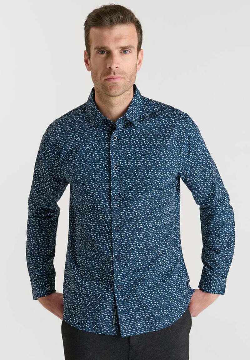 Uomo con capelli corti castani che indossa una camicia a maniche lunghe blu con motivo e pantaloni neri, in piedi con le mani nelle tasche.