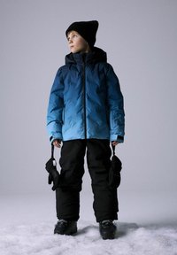 Zils ombre formas pūkainis mētelis, melni sniega bikses, melna beanie cepure un cimdi. Tekstūrēta auduma, rāvējslēdzēji un spiedpogas. Stāv sniegā.