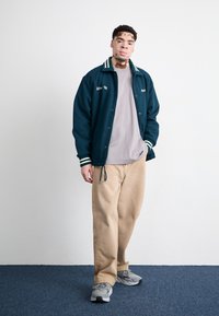 Dark green varsity jacket with white striped collar, gray shirt, beige trousers, and gray sneakers, standing on blue carpet against a white wall.