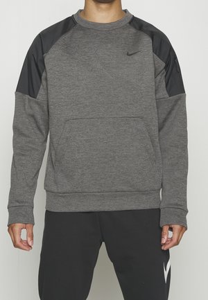 Person trägt einen grauen Nike-Sweatshirt mit schwarzen Schultereinsätzen und schwarze Nike-Hose, steht mit entspannten Armen seitlich am Körper.