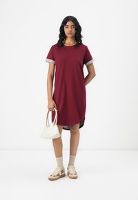 Burgundy t-paita mekko harmailla hihansuilla, polvimittainen, sivutasku. Yhdistetty pieneen valkoiseen käsilaukkuun ja beigeihin sukkiin tennareiden kanssa.
