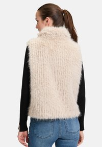 Veste en fausse fourrure de couleur beige clair avec une finition texturée. Dotée d'un col montant et portée par-dessus un haut à manches longues noir et un jean bleu.