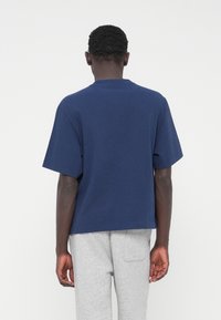 Μπλε ναυτικό βαμβακερό T-shirt με κοντά μανίκια, στρογγυλή λαιμόκοψη και άνετη εφαρμογή. Η πίσω όψη παρουσιάζει έναν απλό, χωρίς διακοσμήσεις σχεδιασμό. Γκρι παντελόνια φαίνονται από κάτω.