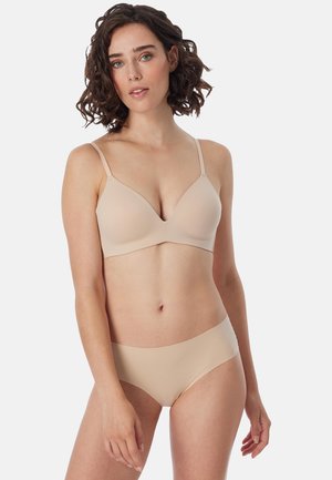 Woman with short curly hair wears matching nude bra and seamless underwear, standing against a plain white background.