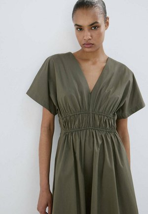 Mujer con el pelo recogido, vestida con un vestido verde oliva con escote en V profundo y cintura fruncida, de pie contra una pared lisa y clara.