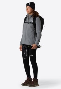 Donna in abbigliamento da escursionismo con zaino nero, giacca in pile grigia, leggings neri, berretto e stivali tiene in mano bastoncini da trekking piegati.