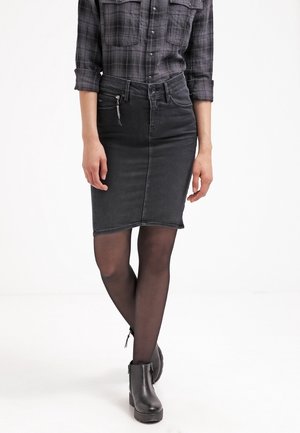 Jupe en denim noir avec une coupe slim, présentant une fermeture éclair à l'avant, une texture lisse et un ourlet brut. Associée à des bottines noires et des collants transparents.