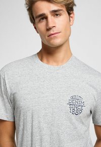 Camiseta de algodón gris de manga corta, con un diseño de texto impreso en azul oscuro en el pecho que dice "DENIM DE CALIDAD DESDE 1889."