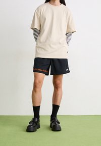 Osoba stojící na zelené podlaze, oblečená v béžovém tričku s krátkým rukávem přes pruhované dlouh�é rukávy, černých šortkách Nike, černých ponožkách a černých botách.