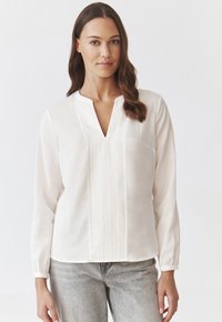 Hvid, langærmet bluse med V-hals og plisseret frontdetalje. Blødt stof og en enkel brystlomme. Båret med lysegrå jeans.