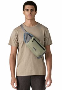 Riñonera verde y gris con un bolsillo delantero con cremallera, material acolchado y correa ajustable, llevada cruzada. De tamaño mediano y forma redondeada.