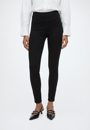 Femme portant un pantalon noir ajusté, une chemise blanche boutonnée et des talons noirs à bout pointu avec deux brides et petits talons.