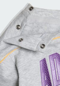 Szara stójka bluzy z metalowymi zatrzaskami, teksturowaną tkaniną oraz fioletowym nadrukiem z motywem kropek. Widoczna żółta linia akcentująca.