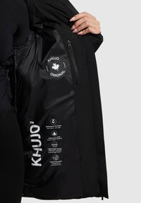 Interno di giacca nera caratterizzato da un tessuto liscio e lucido con marchio bianco, tasca con zip e dettagli impermeabili/anti vento sull'etichetta.