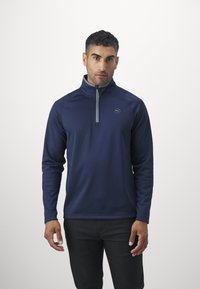Maglione navy con colletto alto e zip a quarto, caratterizzato da un tessuto testurizzato e un logo discreto sul petto, abbinato a pantaloni scuri.