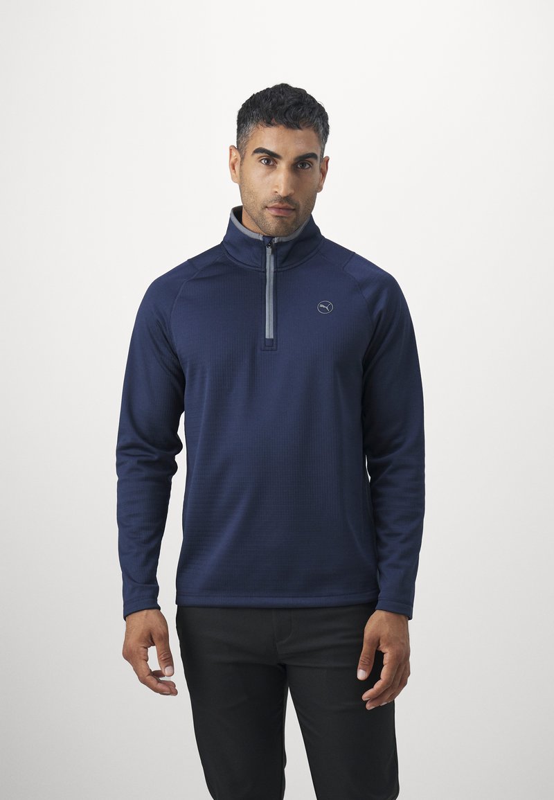 Maglione navy con colletto alto e zip a quarto, caratterizzato da un tessuto testurizzato e un logo discreto sul petto, abbinato a pantaloni scuri.