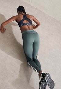 Zwarte sportbeha met uitgesneden rug, groene high-waisted legging met zwarte accenten en witte zijkanten, gedragen tijdens een push-up oefening op een grijze ondergrond.