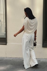Flowy cream blouse draped at shoulders paired with wide-leg white striped pants and black boots, holding a small bag. Simple and elegant design.