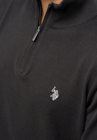 Črni zip-up pulover iz pletene tkanine, z visokim ovratnikom in majhnim srebrnim logo-jem polo na levem prsnem delu.