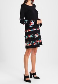 Svart klänning med långa klockärmar; har färgglada blommiga remsor och lager av volanger. Bärs med svarta klackskor med blockklack.