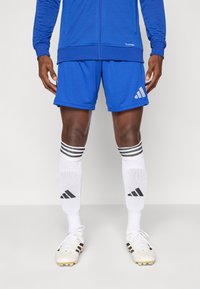 Μπλε αθλητικό τζάκετ με φερμουάρ και σορτς, σετ με λευκές κάλτσες που έχουν μαύρες ρίγες και ποδοσφαιρικά παπούτσια με μαύρες λεπτομέρειες.