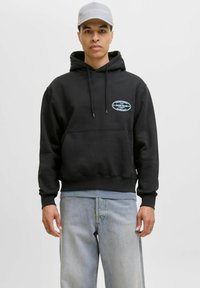 Sudadera negra con un bolsillo frontal, cordones ajustables y un parche gráfico azul. Combinada con pantalones de mezclilla azul claro y una gorra gris.