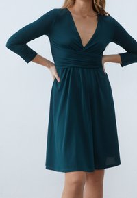 Teal omslagsklänning med V-halsringning, trekvartslånga ärmar och en samlad kjol. Tillverkad av ett slätt, stretchigt tyg med lätt glans.