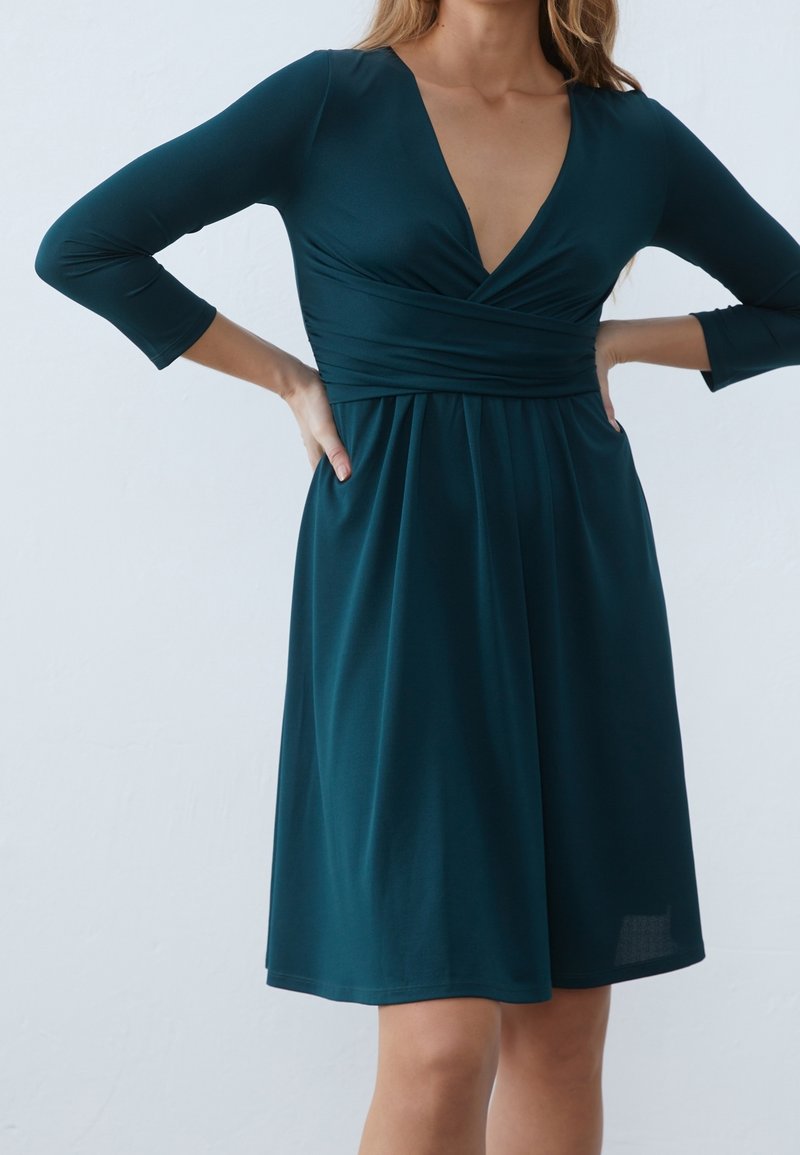 Teal omslagsklänning med V-halsringning, trekvartslånga ärmar och en samlad kjol. Tillverkad av ett slätt, stretchigt tyg med lätt glans.